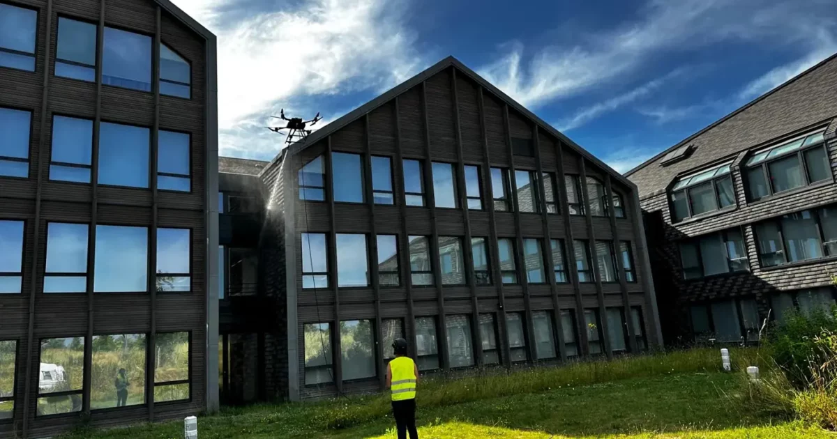 Télépilote en gilet jaune pilotant un drone d'inspection devant la façade vitrée d'un bâtiment pendant la formation Visualiser et Inspecter en drone
